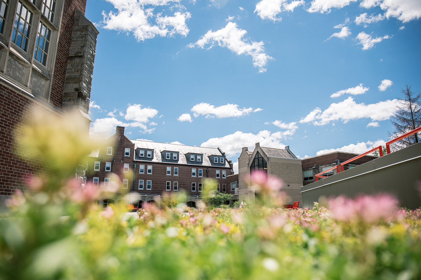 A bright and sunny day at Huron University College, showcasing a courtyard framed by red-brick and stone buildings. The foreground features colorful, out-of-focus flowers, adding a vibrant touch to the image. The buildings in the background have multiple windows and steeply pitched roofs. The sky above is clear with a few scattered clouds, creating a picturesque and welcoming atmosphere.