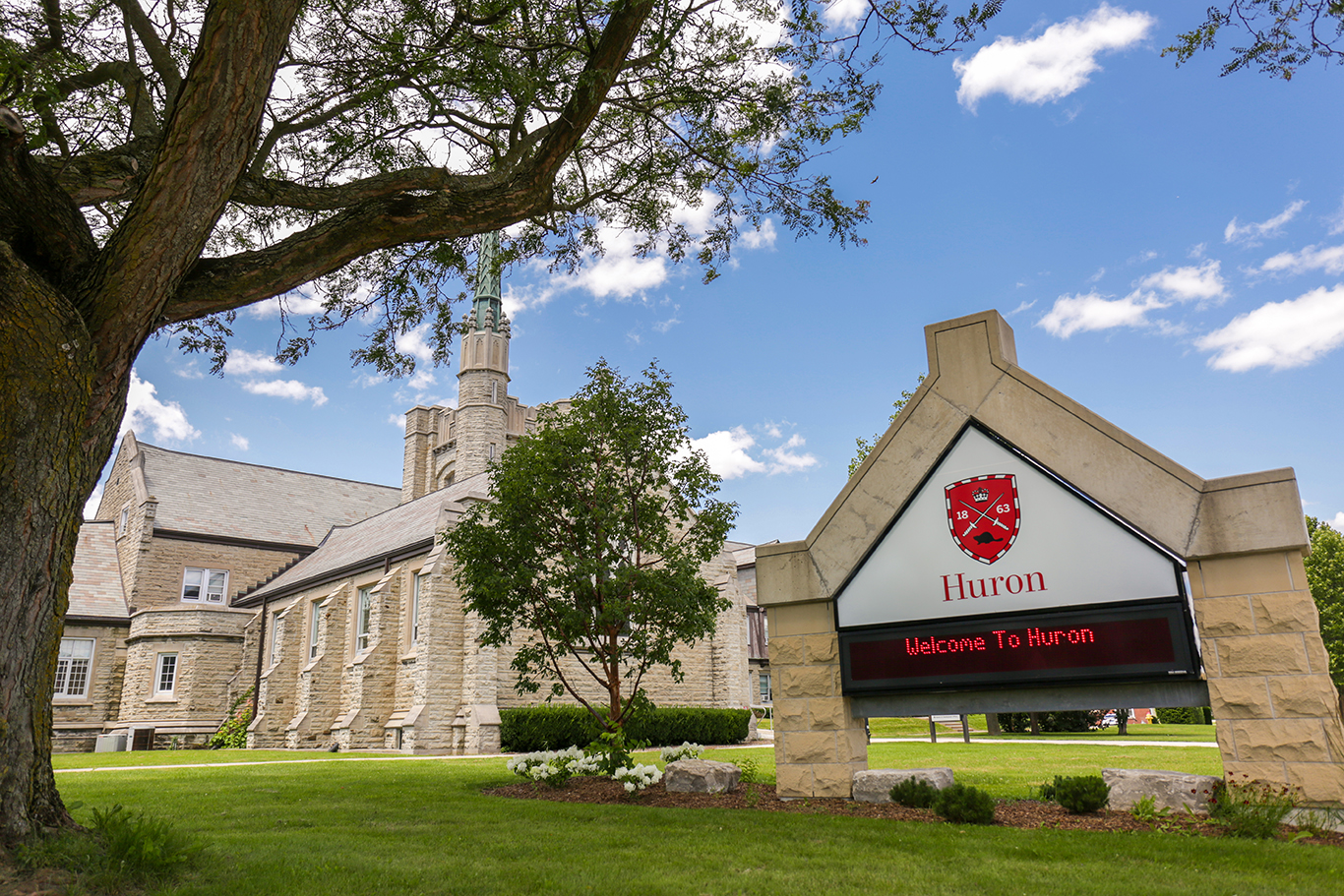 Exterior welcome sign_72dpi A scenic view of Huron University College featuring its historic stone building under a blue sky with scattered clouds. A large tree frames the left side of the image, while a well-maintained lawn and bushes enhance the foreground. On the right, there is a prominent sign with the Huron crest and the words "Welcome To Huron" displayed in red.