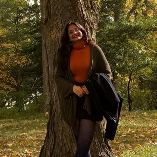 A photo of Anmol Dutta, a researcher who assists in the development of the Courtesans of India project. She is standing outside and smiling on a sunny day, wearing cozy fall clothes.