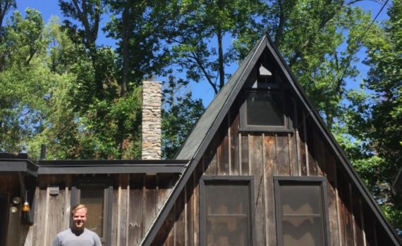 CURL Travel Bursary Winner Andrew French stands outside of Canadian poet Al Purdy's famous A-frame cabin. It's a dark brown, weathered cabin in the middle of the woods.
