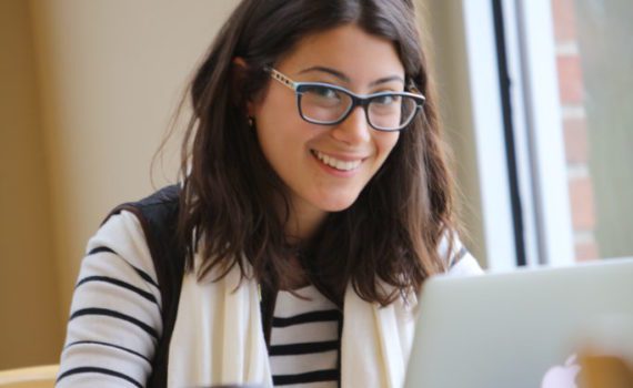 A smiling student with dark hair works at her laptop in the Huron library. She is wearing glasses, a long-sleeved shirt, and a scarf.