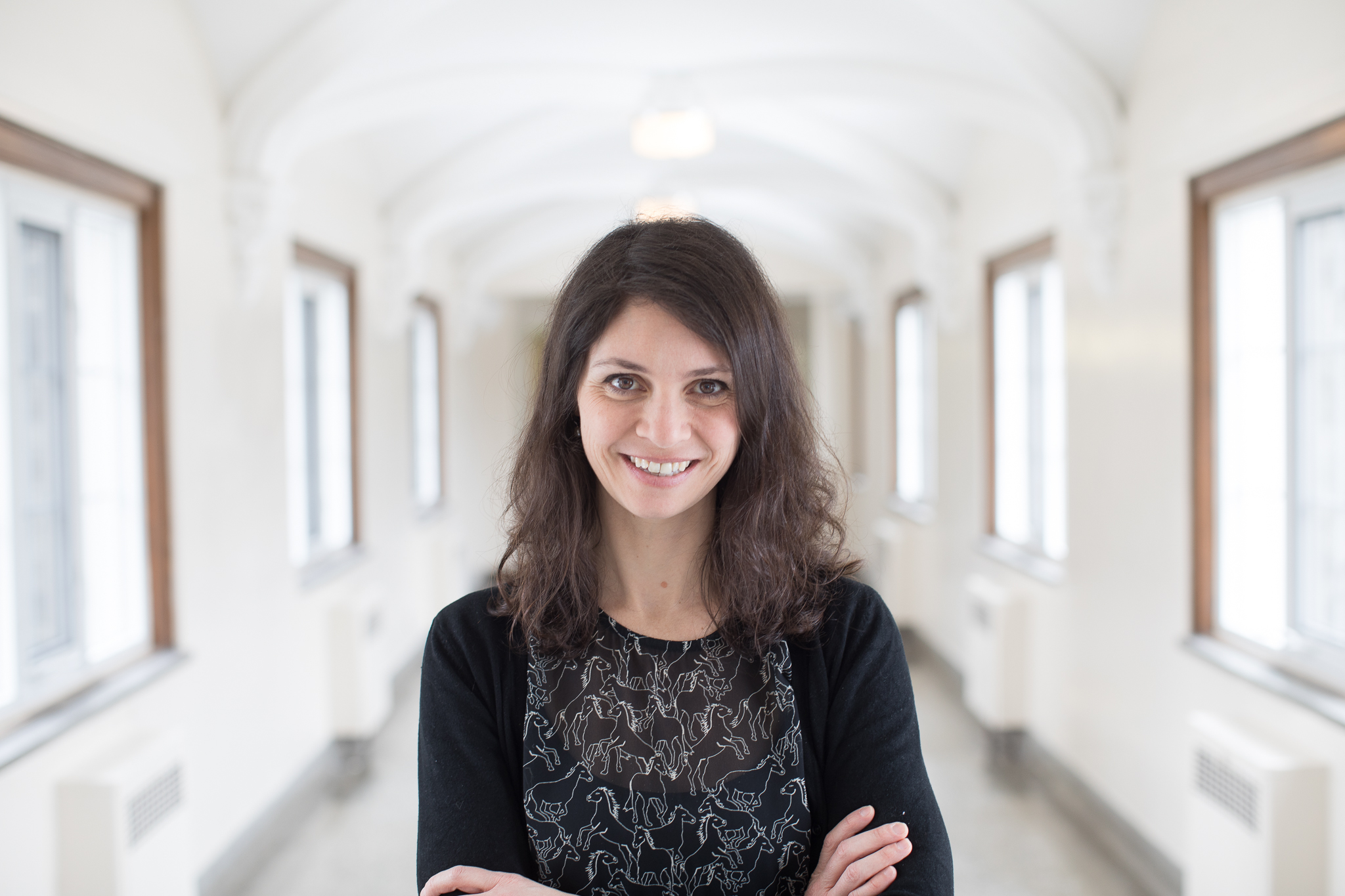A bright, smiling headshot of Dr. Kate Lawless, CGS professor. She has brown, wavy hair and is wearing a dark dress. She is standing in a hallway at Huron University College.