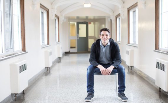 Dr. Michael Kottelenberg sits in a bright hallway at Huron, smiling. He wears a hoodie, jeans, and a denim button-up. He has light skin and short, dark hair.