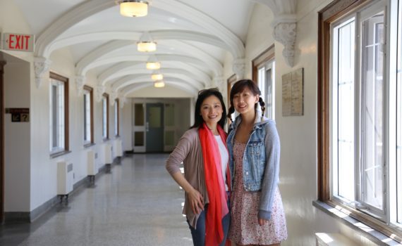 Dr. Lifang (Lucy) He stands in the Huron upper-level hallway, smiling alongside one of her students. Dr. Lifang wears a brown cardigan and a bright red scarf. The student is wearing a jean jacket.