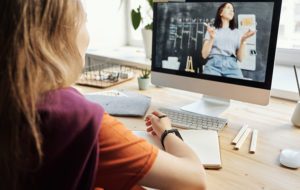 Photo showing the back of a woman watching a video on her computer at home. The video features a woman teaching in front of a chalkboard. Photo by Julia M Cameron from pexels.com.