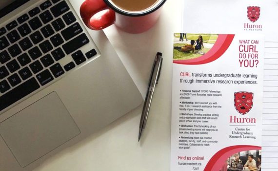 A POV photo of a person's desk. On the left is a laptop. To the right of the laptop is a Huron mug containing coffee. Below and to the right of the coffee are a black pen and a stack of CURL-branded advertisements.