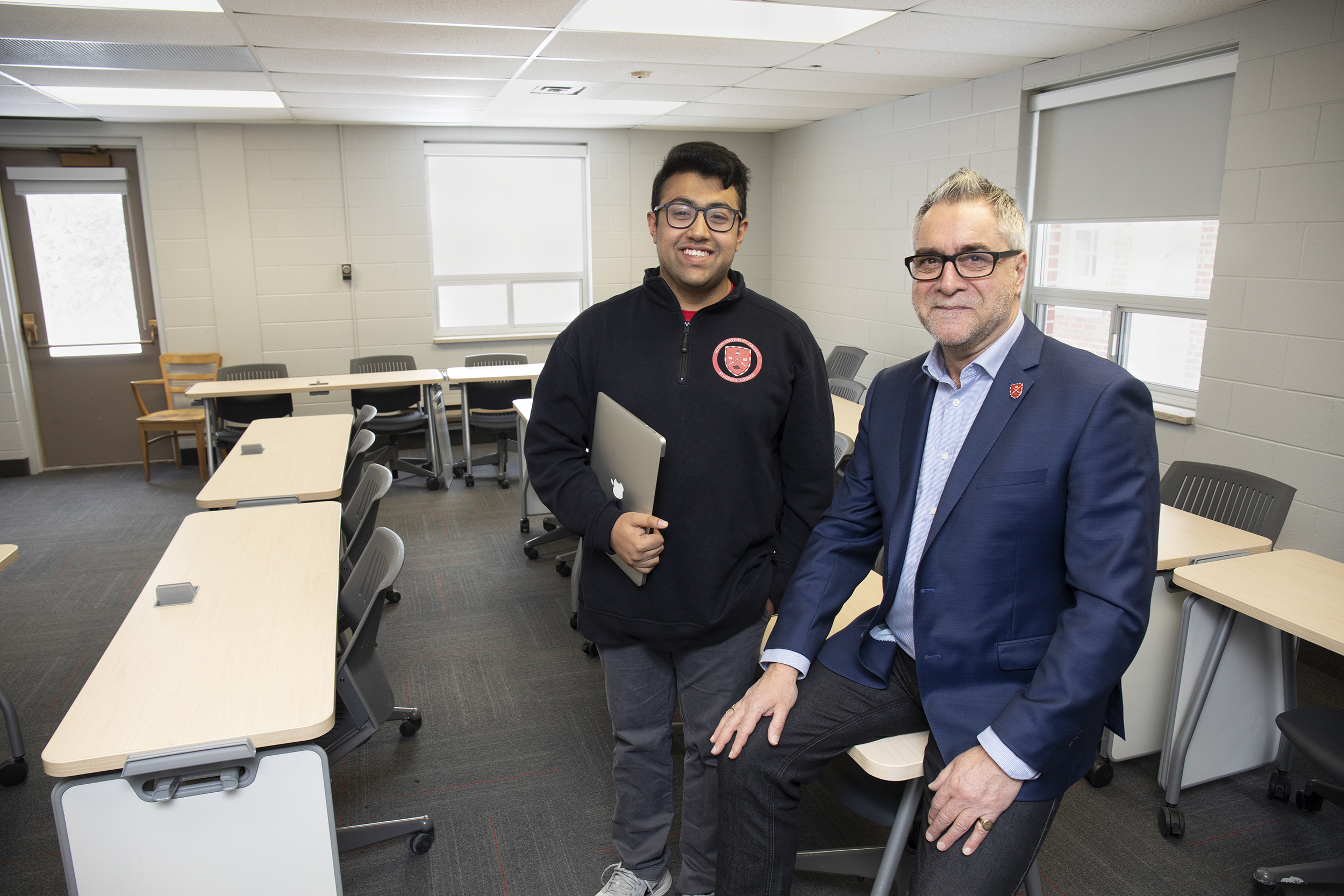 A Huron student in his mid-twenties stands next to an older Huron professor, who is sitting casually on a desk. They are both smiling at the camera.