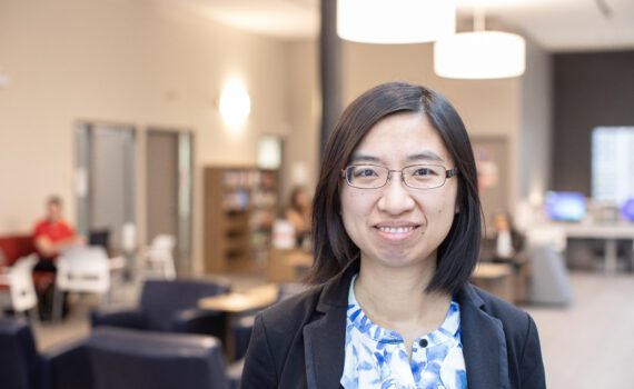 A smiling photo of Dr. Yan Lu. She has shoulder-length dark hair and a blue blazer on, and she's standing in the Huron Library Commons.