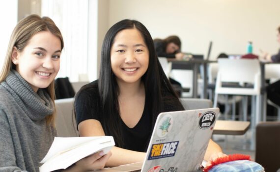 Two Huron students with long hair sit at a table in the Library. They are working on their homework and smiling.