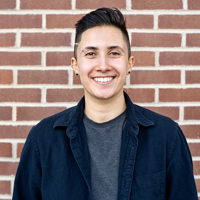 A smiling photo of Cogie Cogan. They have light skin and short dark hair and are standing in front of a clean brick wall. They're wearing a casual button-up shirt over a t-shirt. Their smile is super friendly and endearing.