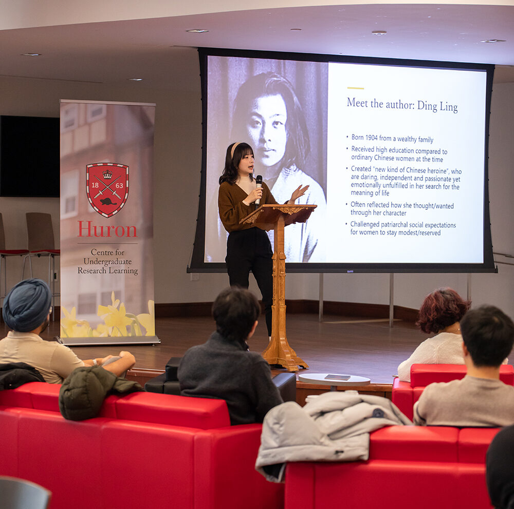 A student with long, brown hair presents her work at the 2023 CURL Fall Exhibition. She is holding a microphone and standing next to a large projector screen. The Beaver Dam is full of students and professors learning from her.