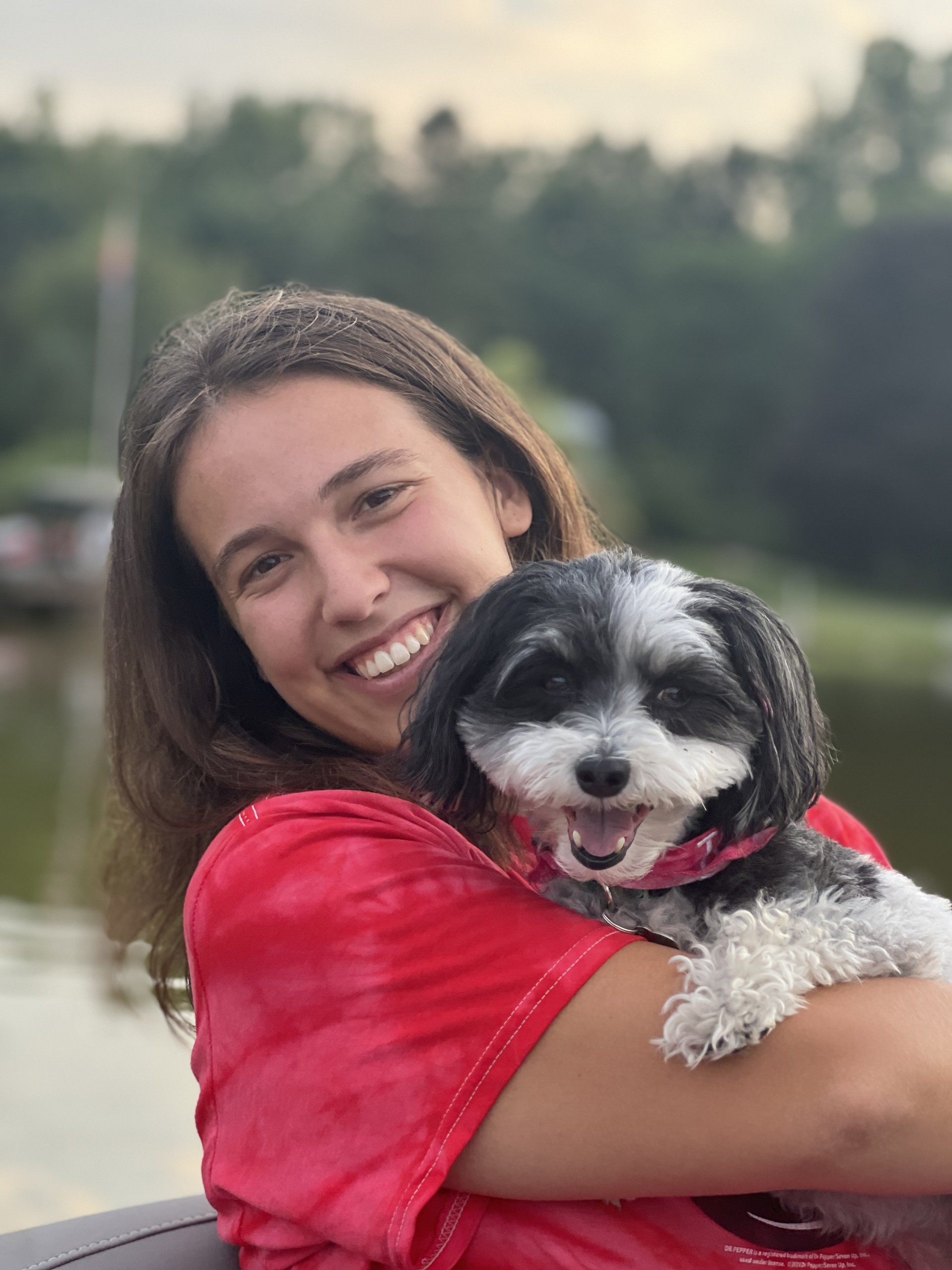 Brianne Headshot A smiling photo of CURL Fellow Brianne Machan holding an adorable black and white dog. She has long, dark hair and a comfy red t-shirt on, and she is standing outside near a lake.