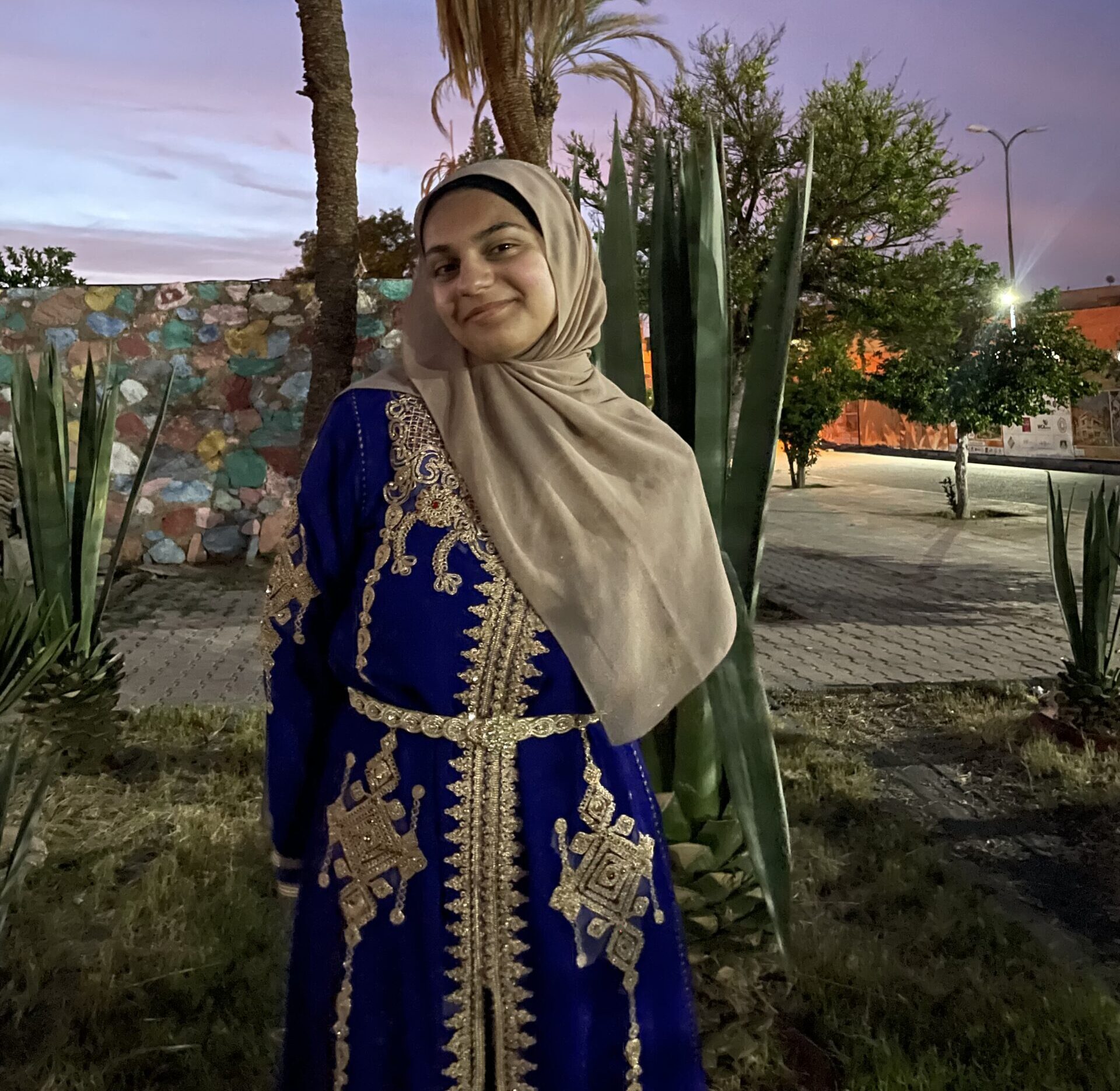 A photo of CURL Fellow Tala El Alam smiling at dusk. She wears an ornate dress with gold detailing and a tan hijab.