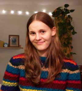 A smiling photo of CURL Fellow Julia Hoytema. She wears a comfy-cozy knit sweater with colourful stripes and has long, straight, auburn hair.